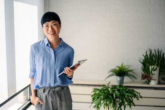 Asian woman in blue shirt holding a tablet and smiling indoors near large windows and plants, looking confident and approachable like a professional