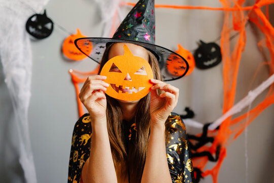 A person wears a spooky witch hat and holds a bright pumpkin mask while surrounded by vibrant Halloween decorations and cobwebs