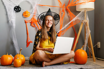 A girl wears a playful witch costume, smiling while using her laptop surrounded by pumpkins and Halloween decorations in a cozy space