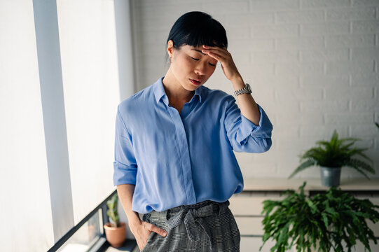 Tired Asian woman standing indoors with hand on forehead, expressing stress, fatigue, or headache, representing exhaustion and emotional struggle