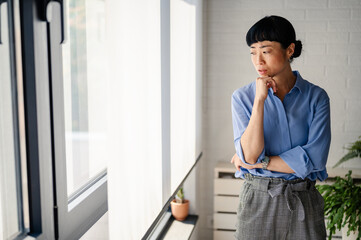 Worried middle-aged Asian woman standing by the window with hand on chin, looking outside thoughtfully, expressing concern, doubt, or contemplation