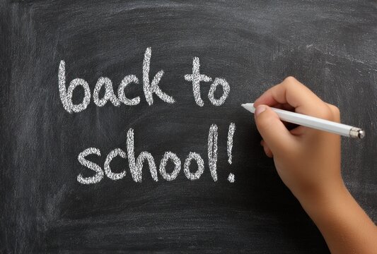 A child's hand writes "back to school!" on a dark chalkboard with white chalk, conveying a sense of excitement and education