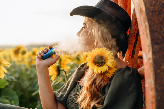 Woman enjoying a relaxing moment in a sunflower field while vaping and wearing a wide-brimmed hat