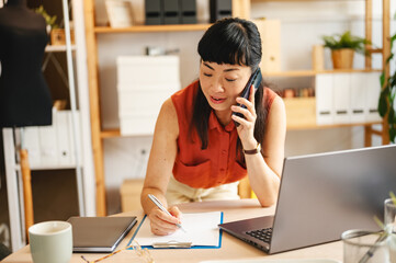Small business entrepreneur in a modern studio office talking on phone and writing notes at desk with laptop, planning and managing work tasks