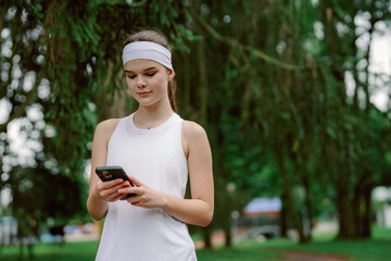 A young woman in athletic wear stands in a lush green park, focused on her smartphone. The setting is calm, with trees surrounding her, suggesting an active and leisurely lifestyle. © m-art