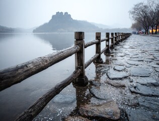 Serene lakeside scene with a weathered wooden fence lining a cobblestone path, leading towards a distant, misty mountain range on a cloudy day