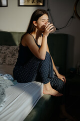 A young woman sits on the edge of her bed, sipping from a glass while basking in the soft morning...