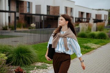 A woman walks confidently along a paved path in a contemporary residential area. She wears a stylish outfit with a sweater tied around her shoulders and carries a bag.