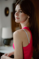 Fototapeta premium A young woman with long brown hair is standing casually in a bright indoor space. She wears a pink top and looks over her shoulder with a confident expression, enjoying a moment of relaxation.