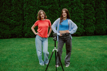 Two young women stand confidently in a green garden, striking playful poses in casual outfits while...