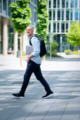 Confident middle-aged man walking along a city street and holding laptop in his hand