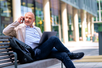 Businessman relaxing on a bench in a city environmental and using laptop and smartphone