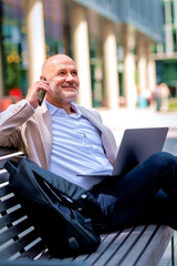 Businessman relaxing on a bench in a city environmental and using laptop and smartphone