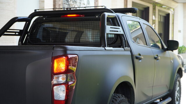 Rear three-quarter view of a modern matte black double-cab pickup truck with customized off-road accessories. The vehicle features a black roll bar, roof racks, and illuminated red tail lights.