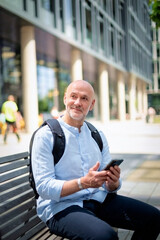 Confident middle aged man sitting on a bench on the city street and using smartphone