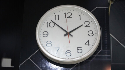 round wall clock with white face, black numbers, black hour and minute hands, red second hand showing time, mounted on dark patterned wall with visible wire