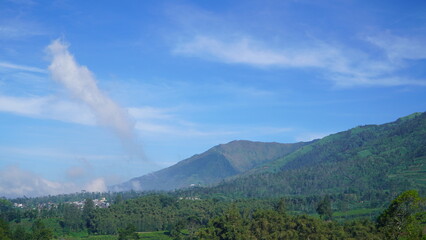 lush mountain landscape under vivid blue sky, dense greenery and scattered mist near village base create serene contrast across layered terrain and soft cloud formations