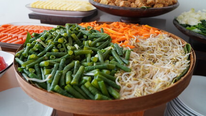 wooden bowls filled with fresh vegetables including green beans, bean sprouts, julienned carrots, chopped lettuce, tofu, and pineapple in colorful buffet or salad bar setup