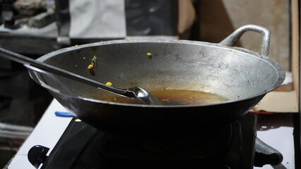 large metal wok on black stovetop with ladle and liquid containing food remnants, possibly corn kernels, in kitchen setting with white surfaces and background items