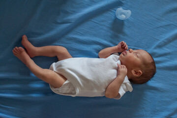 A newborn baby in a white onesie sleeps soundly on a blue sheet. A light blue pacifier rests next to the baby. The baby is peacefully asleep.