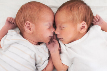 Two newborn twin brothers lie side by side, fast asleep. Their heads are touching, and their hands meet in the middle, forming a heart shape.