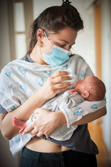 Young woman wearing a surgical mask holds and feeds newborn baby with a bottle. The baby is wrapped in a blanket. This intimate moment occurs during the day inside of the home.