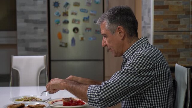 Middle-aged Man in a Plaid Shirt is Sitting at a Dining Table, Eating Grilled Chicken, Grilled Zucchini, and Tomato Salad. A 55-year-old, gray-haired man enjoying lunch in the peace of his apartment.