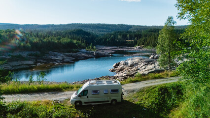 Nestled in the heart of Norway, a cozy camper van sits near a calm blue lake, surrounded by verdant forests and rocky outcrops under a brilliant sky. Perfect for adventure seekers. © Fokke Baarssen