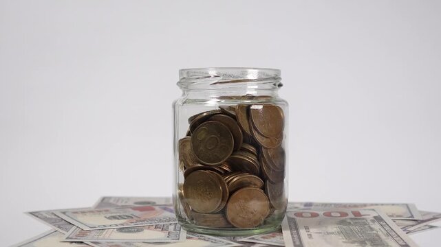 Shiny gold coins fill inside a rotating transparent glass jar, with scattered dollar bills around it, symbolizing wealth accumulation, savings, and financial growth.