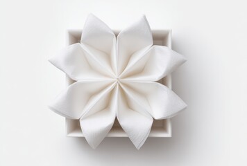 A delicately folded white napkin arranged in a flower-like pattern inside a square dish, resting on a clean, white surface