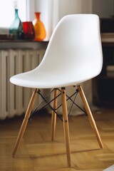 White plastic chair with wooden legs sits on a parquet floor, light streaming from a nearby window with decorative glass vases on the sill