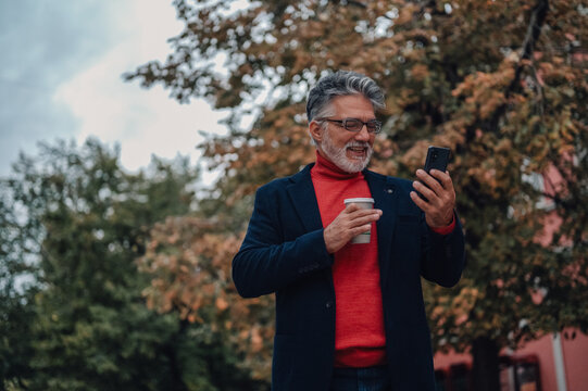 Stylish senior man using smartphone and drinking coffee outdoors in autumn - Powered by Adobe