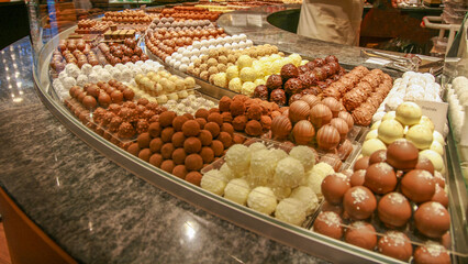 A variety of chocolate truffles and pralines on display in a glass display case in a pastry shop. An assortment of sweet, gourmet desserts including milk, dark and white chocolate.