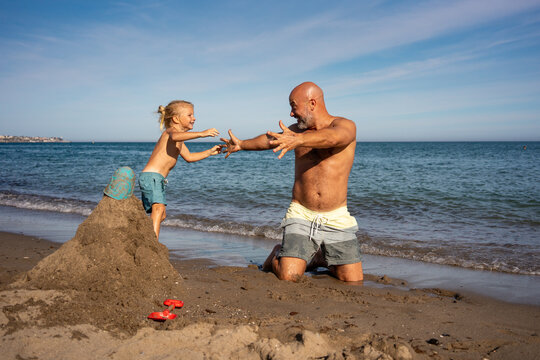 Joyful father and son playing together on the beach near a sandcastle, smiling and reaching for each other with the sea and blue sky in the background