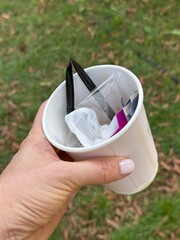 Hand holding paper cup filled with assorted plastic trash over grassy background