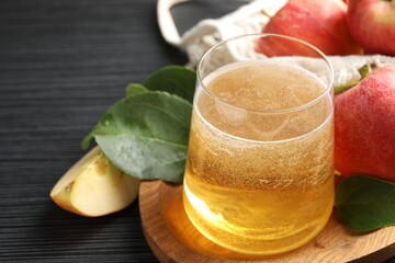 Fresh cider in glass, apples and green leaves on black textured table, closeup