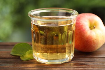Delicious cider in glass, apple and green leaves on wooden table outdoors, closeup
