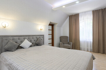 A calm bedroom is shown with a large bed, neutral color scheme, grey armchair, and window with curtains. The room has gentle lighting