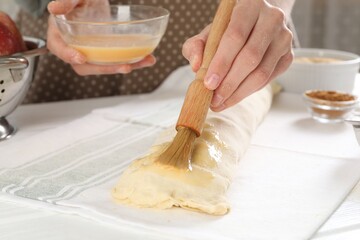 Woman spreading egg yolk onto raw apple strudel at table in kitchen, closeup