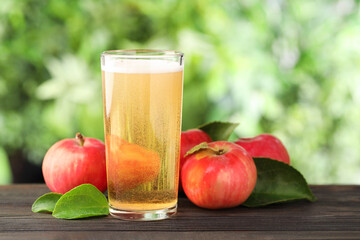 Delicious cider in glass, apples and green leaves on wooden table outdoors