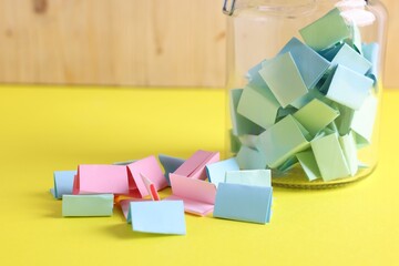 Colorful paper notes in glass jar on yellow table, closeup