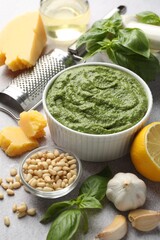 Fresh pesto sauce in bowl and ingredients on light grey table, closeup