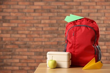 Backpack, paper planes, lunchbox and apple on wooden table against red brick wall. Space for text