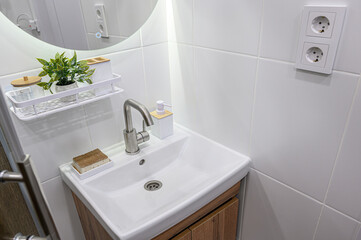 A clean, modern bathroom sink area with white tiles, a round mirror, a stainless steel faucet, and wooden cabinets