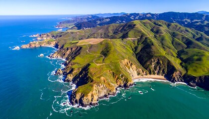 Aerial vista of a rugged coastline with verdant hills meeting the ocean. Winding road visible. Blue sea under a clear sky