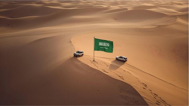 An aerial shot of a white 4x4 vehicle parked on top of a towering sand dune in the vast, serene desert. A Saudi flag flies high from the vehicle, symbolizing exploration, adventure, and national pride