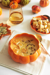 Creamy pumpkin soup with croutons and seeds, served in a pumpkin-shaped bowl on white wooden table.