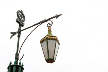 Decorative streetlair in vintage style against background of bright sky. Lantern is made of metal with green and golden trim, suspended on elegant forged structure.