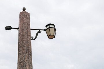 Decorative street lamp in vintage style against the sky. The lantern is made of metal and is suspended on a stone column made of red granite.