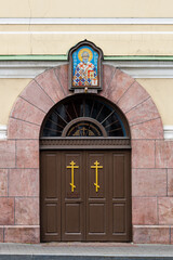 Entrance to Orthodox Church with arched portal, icon of saint and two golden crosses on wooden doors.